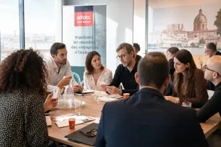 Professional business networking event in a sunlit meeting room, entrepreneurs exchanging contacts and discussing ideas around a table with southern France ambiance.