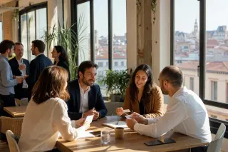 Professionals networking around a communal table in a sunlit coworking space with a southern French city view visible through large windows.