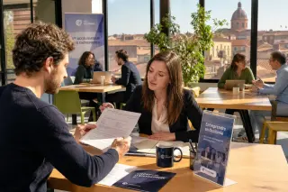 Entrepreneur reviewing documents with advisor in a sunlit French coworking space, with hints of Occitanie architecture visible through the window.