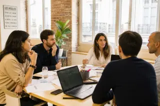 Entrepreneurs in a sunlit, modern French coworking space having a focused planning discussion around a table with papers and laptops.