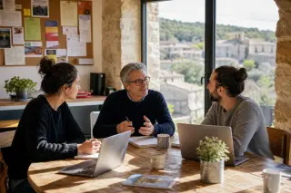 Small group of diverse entrepreneurs in a sunlit coworking space in rural southern France, engaged in a collaborative meeting with laptops and coffee.
