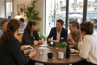 Business owners in casual-professional attire networking in a sunlit coworking space, gathered around a table with Occitanie cityscape visible through large windows.